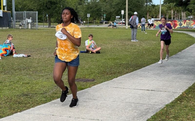 Two girls wearing marathon bibs sprint down a sidewalk while other kids watch from the grass.