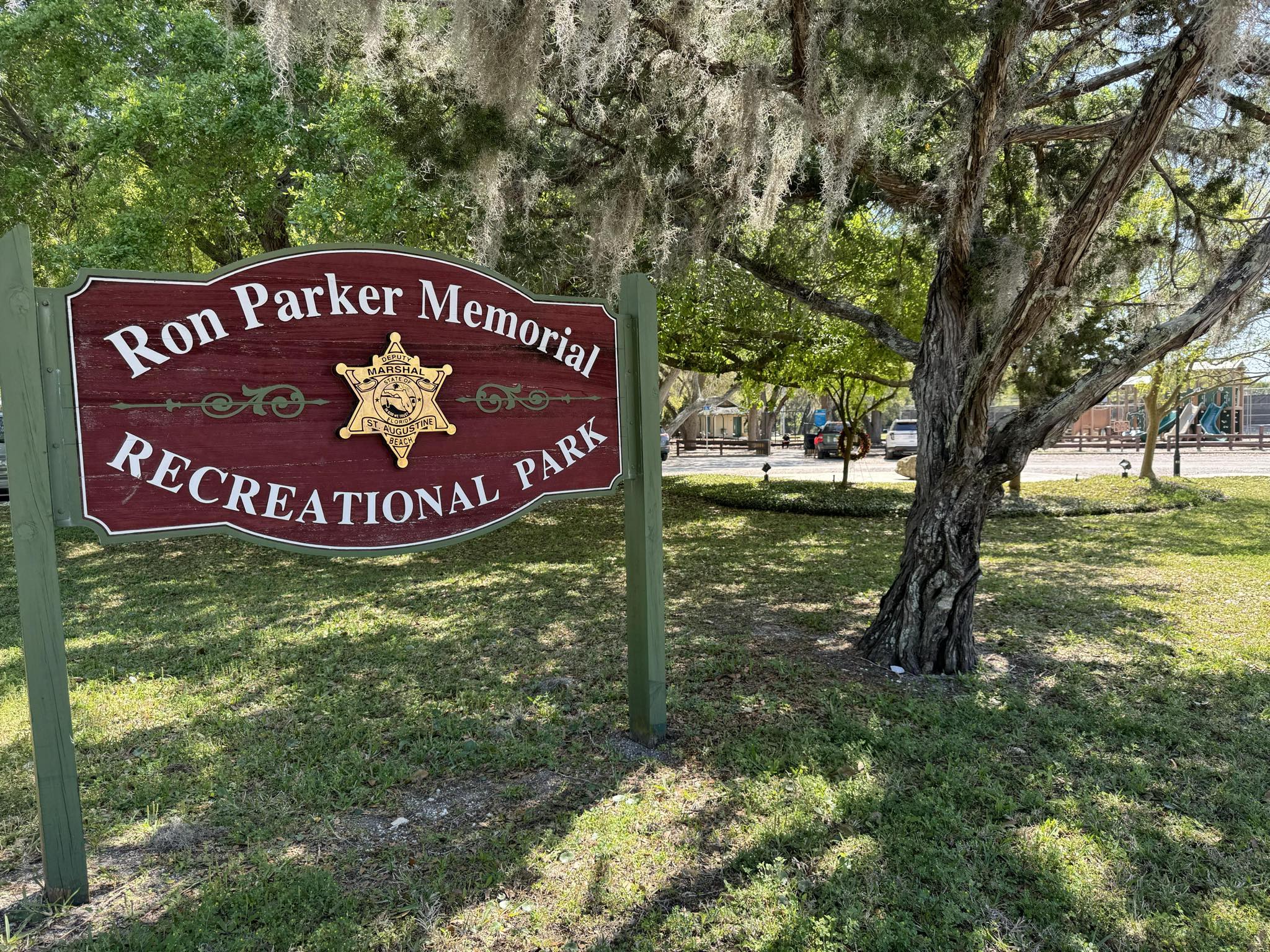 A sign for Ron Parker Park next to a tree, in front of the park's playground