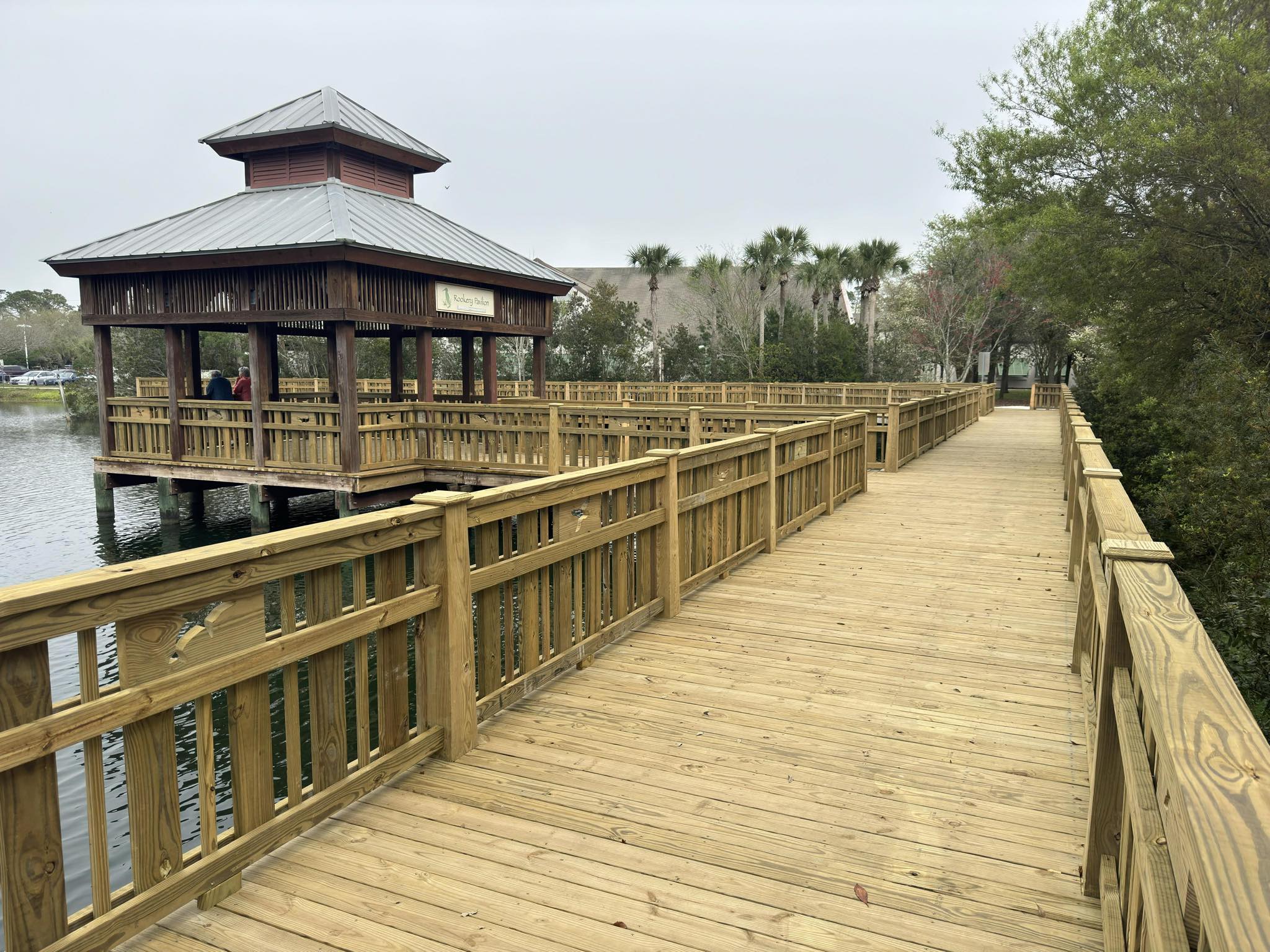 Wooden walkway on a lake surrounded by trees, with a covered pavilion to the left.