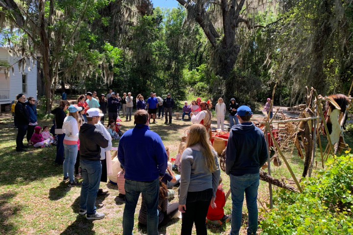 A group of people gather in a wooded clearing in a park to watch a historical demonstrator near a table of artifacts.