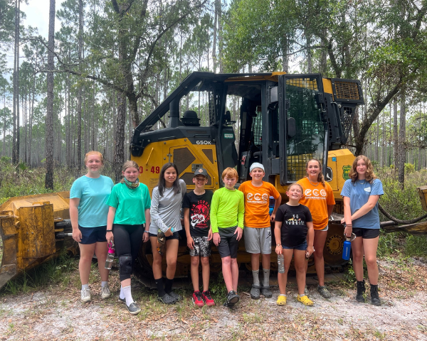 Nine children posing in front of an earth-mover in a forest clearing.