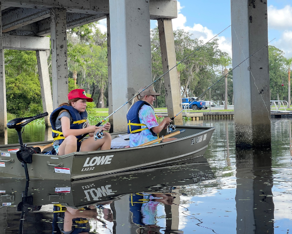 Two girls fish from a motorboat under a bridge.