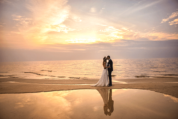 couple dressed in wedding attire standing on sandy beach with sun peeking through clouds