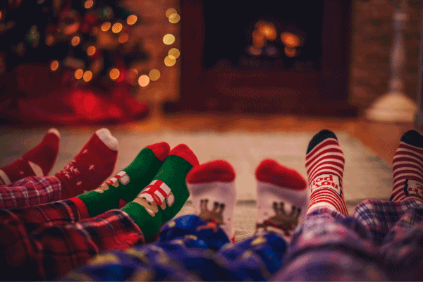 Four pairs of legs and feet in festive pajamas and socks, with a Christmas tree and fireplace in a decorated living room the background