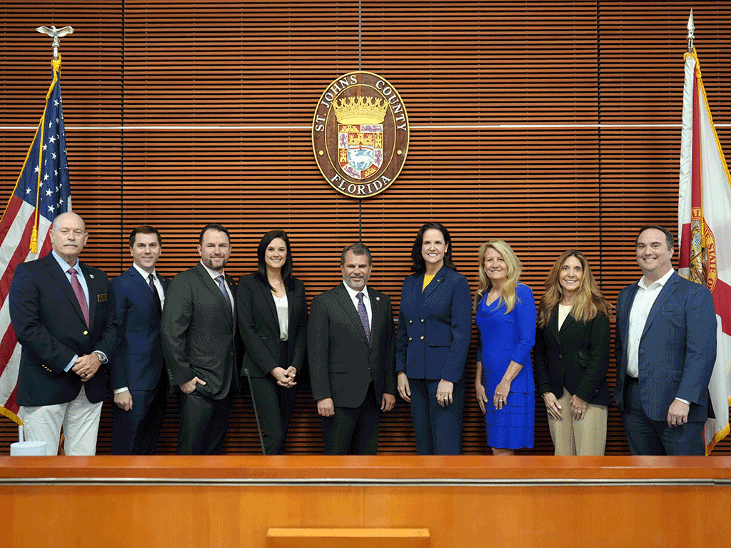 State Representatives and St. Johns County Commissioners gather under the St. Johns County seal and the United States and State of Florida flag in an auditorium