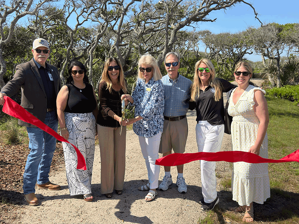 Commissioner Clay Murphy, Parks and Recreation Assistant Director Jamie Baccari, Commissioner Ann Taylor, and Chair Krista Joseph stand with Marcia Mussallem Byles and two members of her family as they cut a red ribbon with a huge pair of golden scissors at the entrance to a forested walking trail.