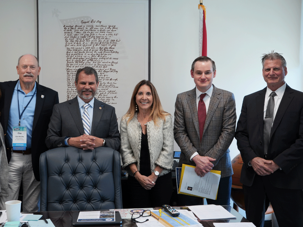 Officials standing behind a desk in an office with a framed picture and the Florida state flag in the background.