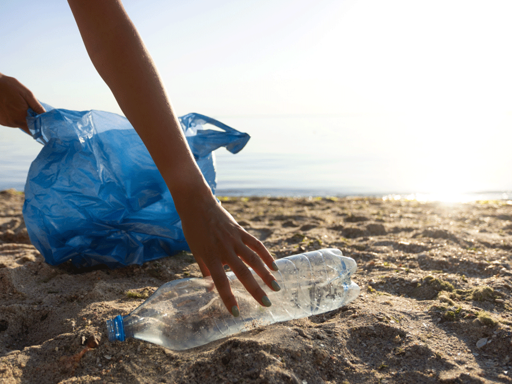 An individual picking up an empty water bottle on the beach and holding a blue plastic bag.