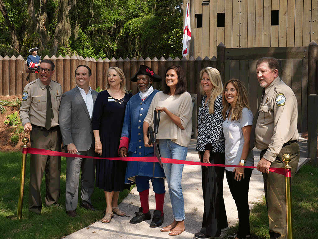 Commissioners Whitehurst, Arnold, Taylor, and Chair Joseph smile behind a red ribbon. Commissioner Arnold holds giant scissors to cut the ribbon.