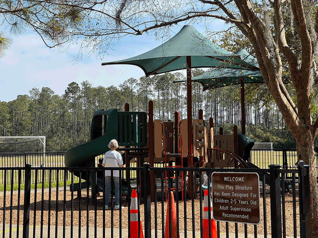 An older woman watches over small children playing around a shaded play structure.