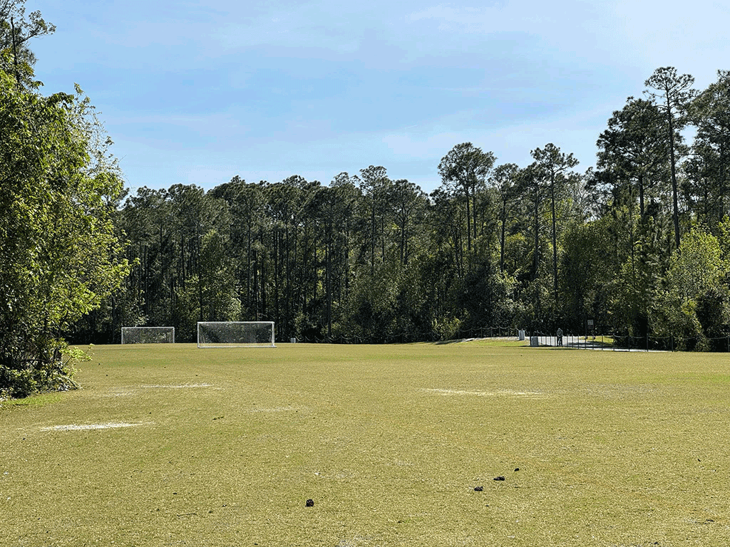 A paved walking/biking path runs around a wide grass field with soccer goals, bordered by the woods.