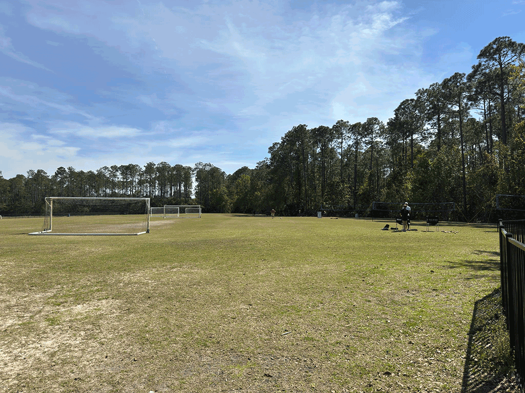 A few people stand in the shade on a grass field with soccer goals and volleyball nets.