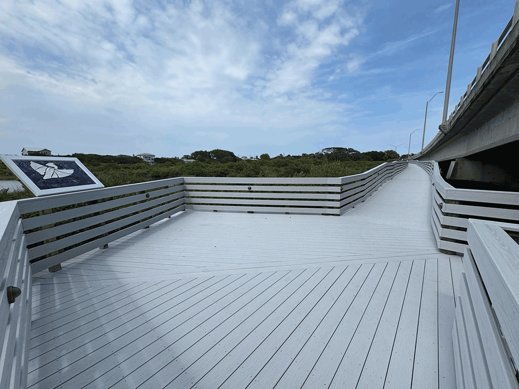 A newly painted wooden boardwalk that runs along an overpass, with a view of the surrounding marsh and scrubland.