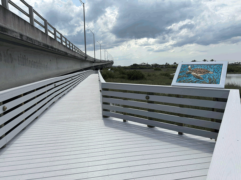 A newly painted wooden boardwalk with a sea turtle mosaic that runs along an overpass, with a view of the surrounding marsh and scrubland.