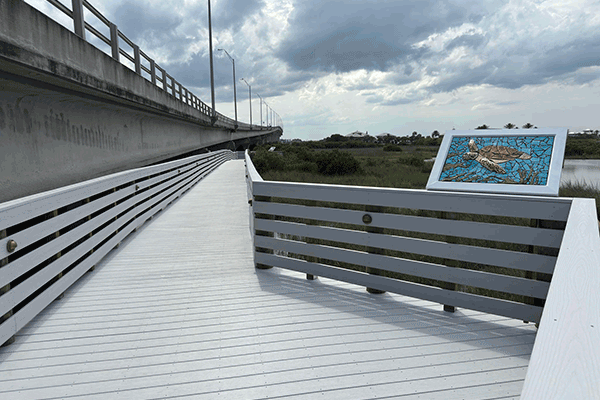 A newly painted wooden boardwalk with a sea turtle mosaic that runs along an overpass, with a view of the surrounding marsh and scrubland.