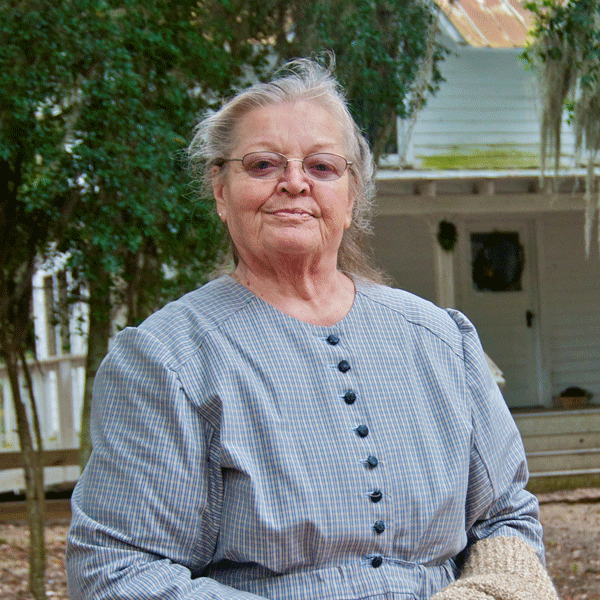 A woman in an antique dress stands outside with a colonial house in the background