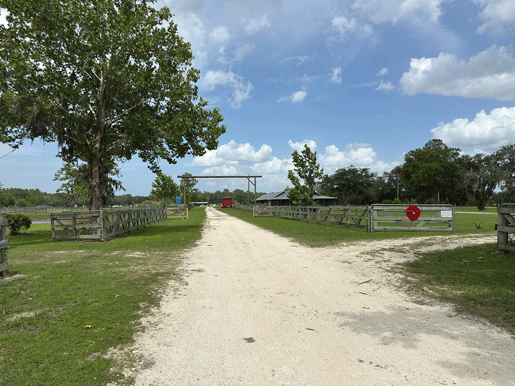 A dirt road leading past fenced pastures through a gate to an equestrian center.