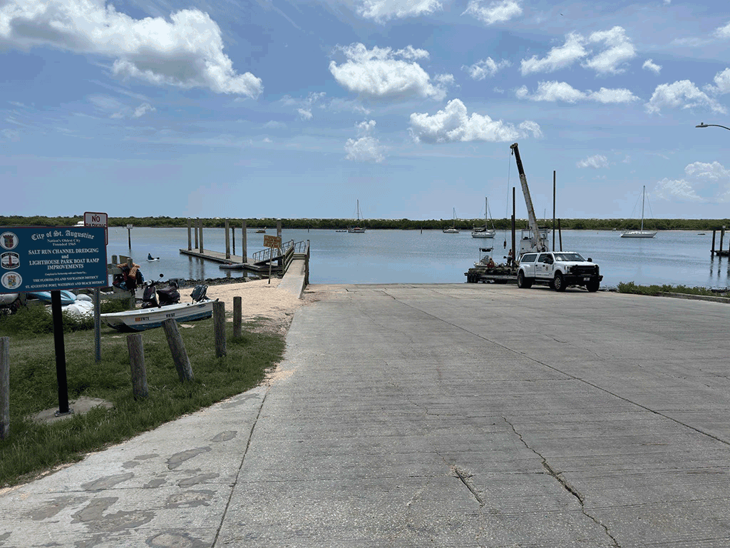 A wide concrete boat ramp with an attached wooden dock and sailing vessels visible in the background.