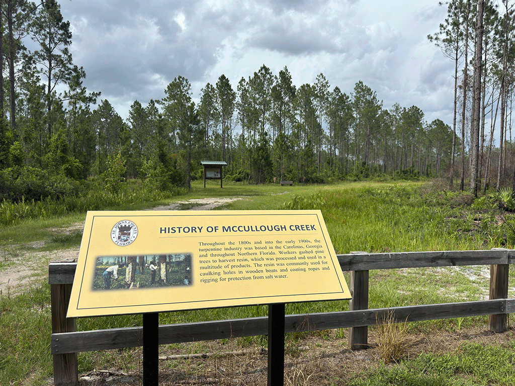 A sign for McCullough Creek East Park next to a gravel path leading to a small parking lot outside a forest trail.