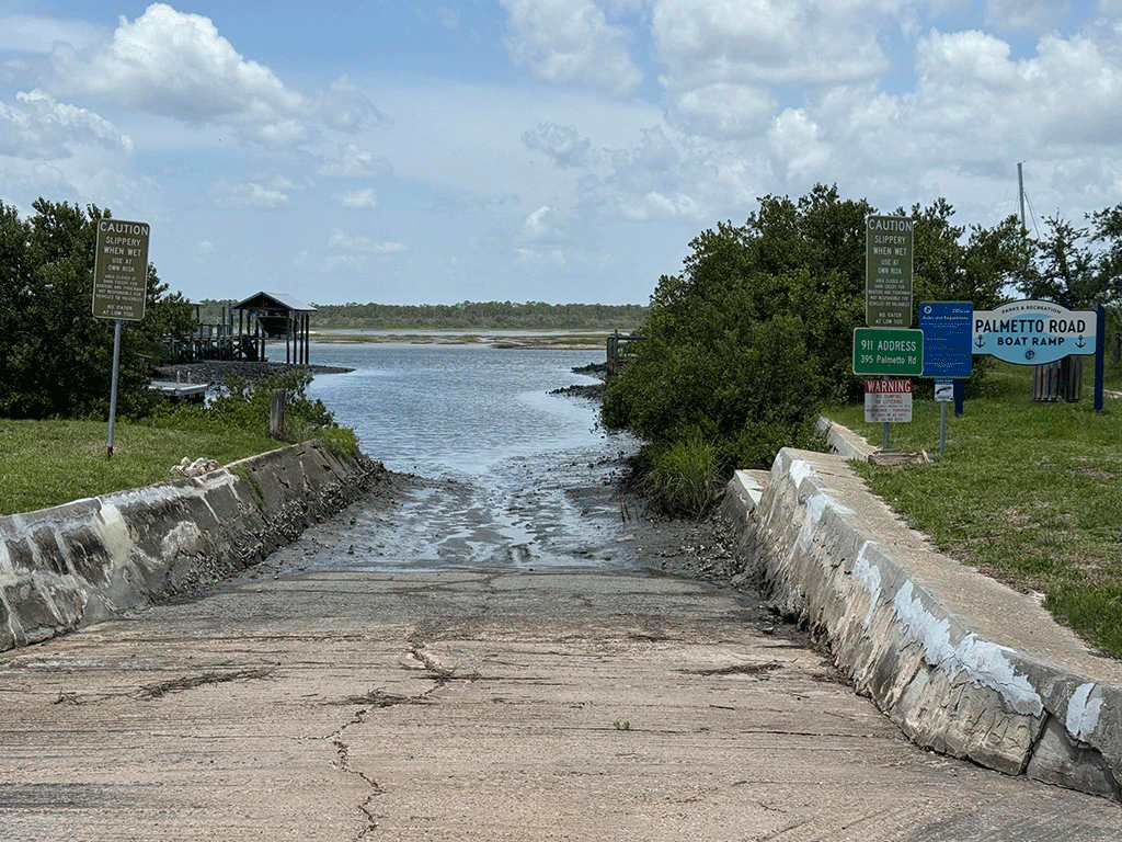 A muddy concrete boat ramp into the Intracoastal, with signs and bushes on either side and a dock visible in the background