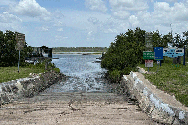A muddy concrete boat ramp into the Intracoastal, with signs and bushes on either side and a dock visible in the background