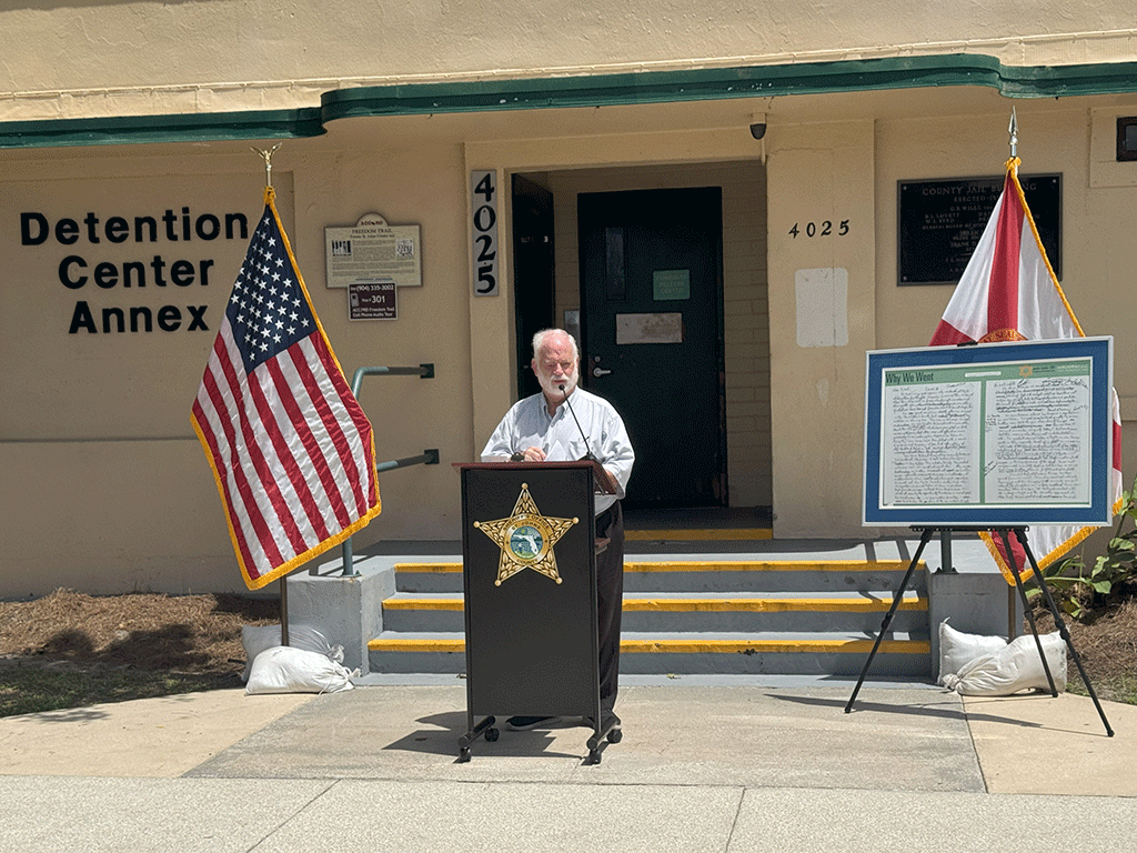 A speaker stands at a podium placed in front of the Detention Center Annex.