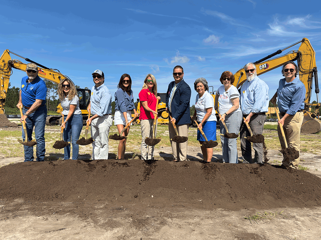 Ten people pose with uplifted golden shovels full of dirt at a construction site, with a pile of dirt in front of them and three construction vehicles behind