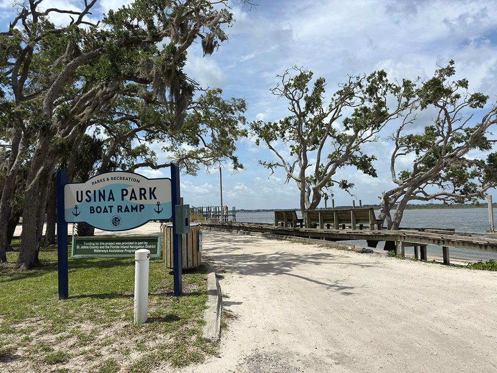 A sign for Usina Park Boat Ramp next to a gravel driveway past a wooden dock