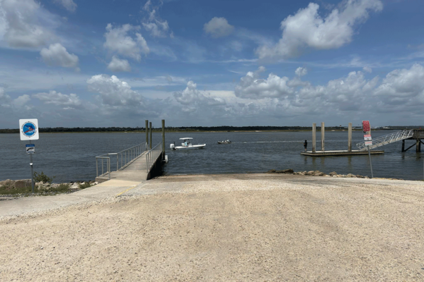 A gravel boat ramp leading into the Intracoastal, with boats passing by and a man fishing off a floating dock in the background.