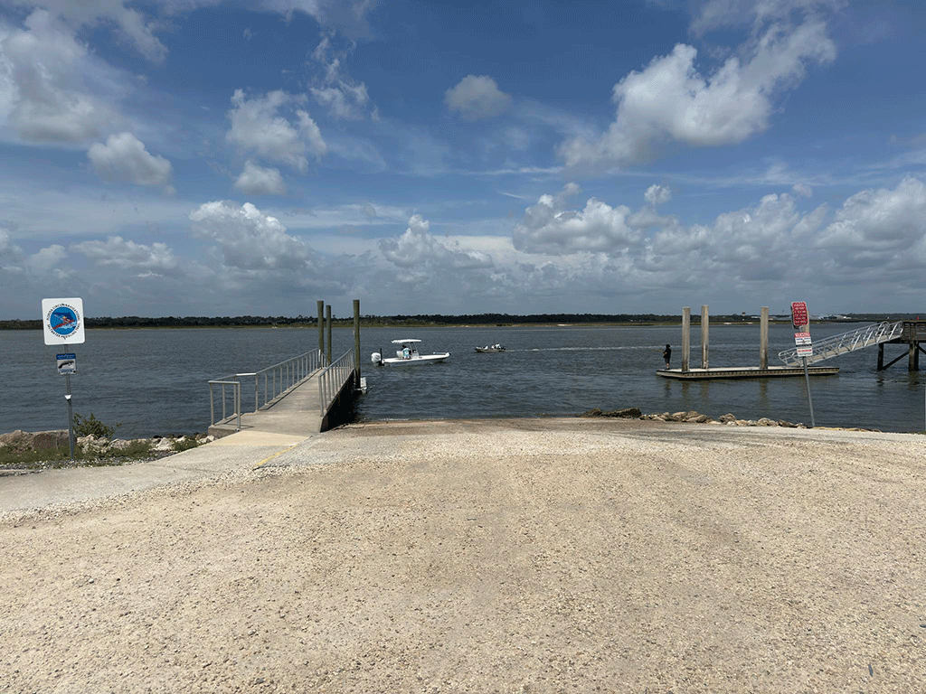 A gravel boat ramp leading into the Intracoastal, with boats passing by and a man fishing off a floating dock in the background.