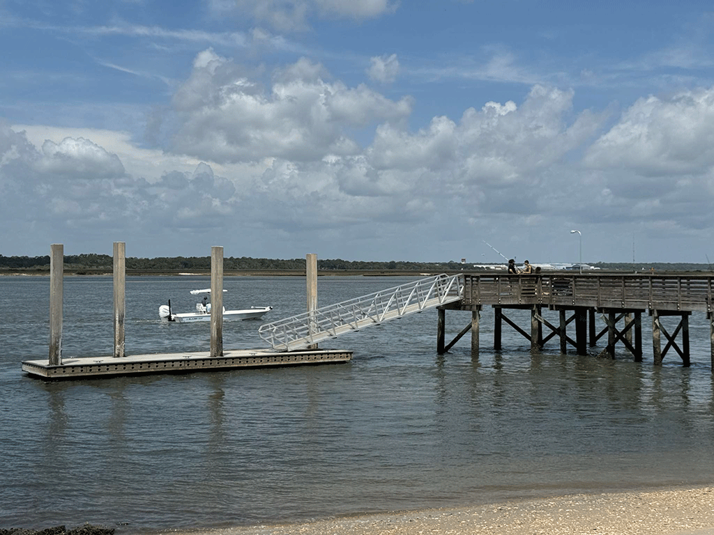 People fish off a wooden pier with an attached floating dock, while a small boat passes in the background.