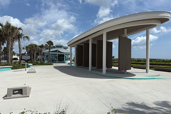 A concrete amphitheater by a beach, with cornhole to one side and a play area and pavilion in the background.