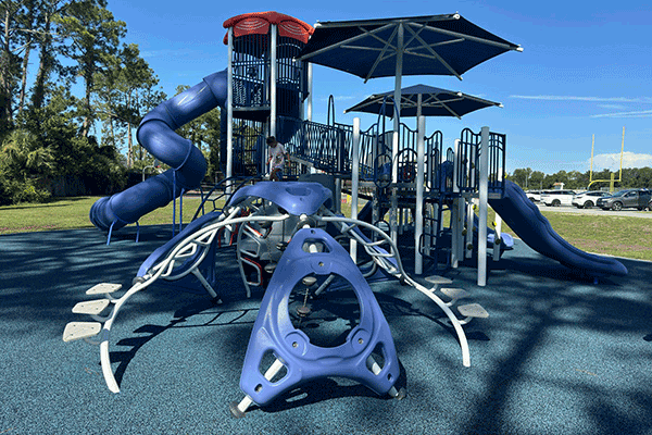 A boy plays on a playground, complete with jungle gym and slides, near a sports field and a parking lot