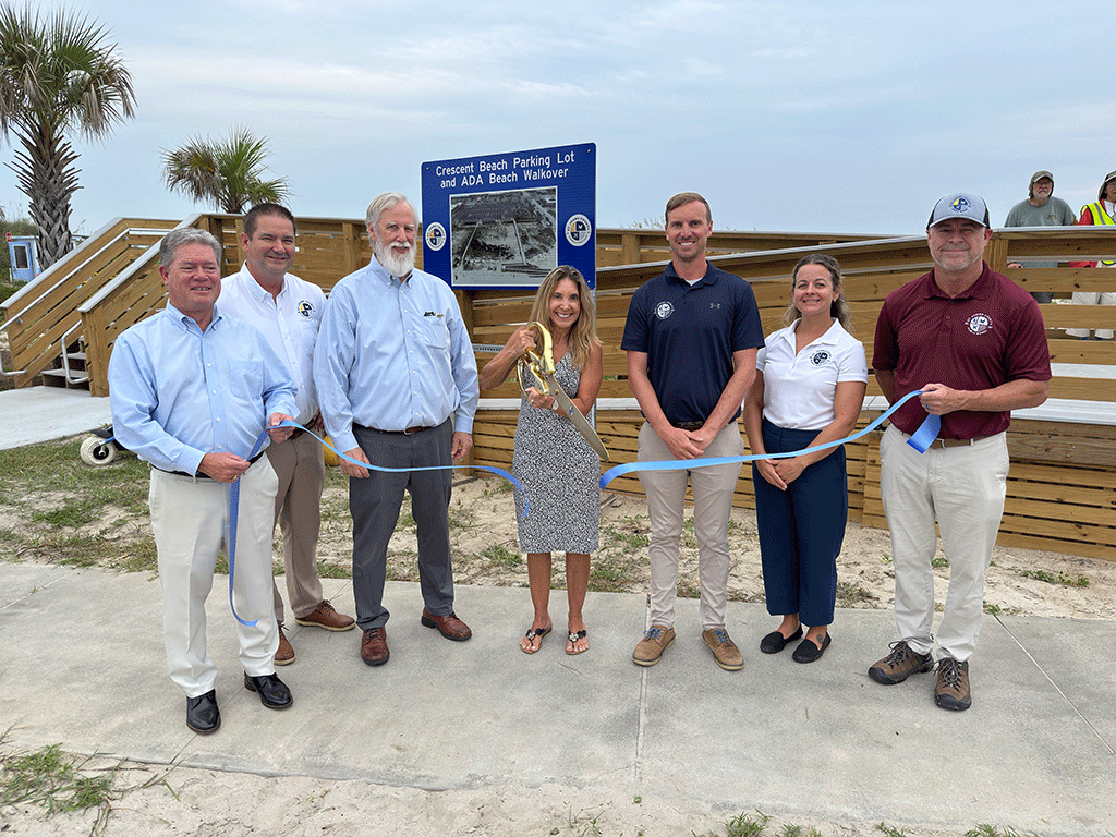 County Engineer Duane Kent, Parks and Recreation Director Ryan Kane, Deputy County Administrator Brad Bradley, Natural Resources Superintendent Nathan Otter, Beach Operations Manager Sara Perez, and CIP Manager Lance Alred stand behind a blue ribbon, with a beach walkover behind them, while District 5 Commissioner Ann Taylor cuts the ribbon with huge golden scissors.