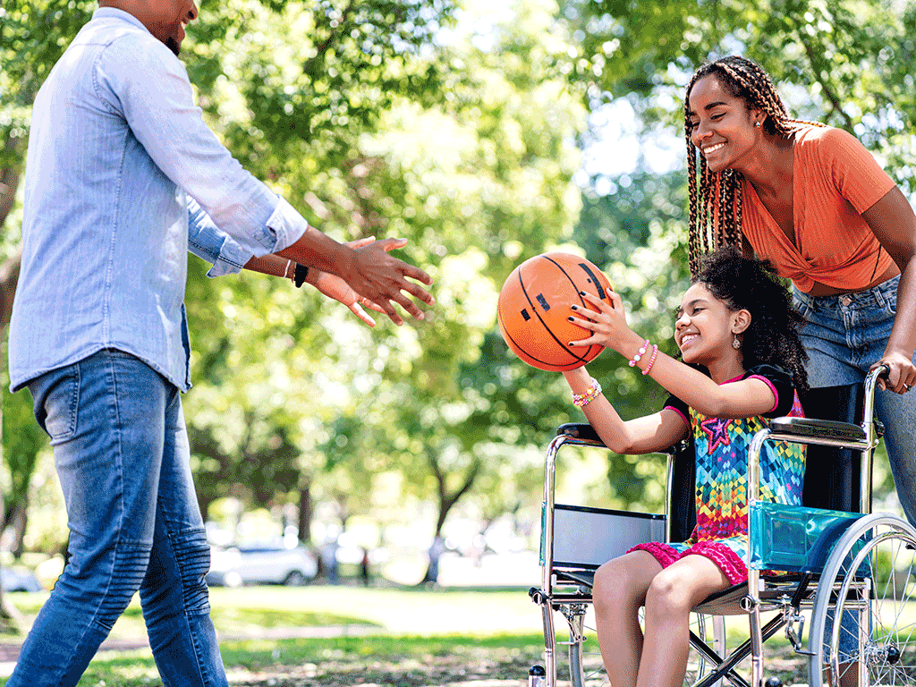 A little girl in a wheelchair plays basketball with two adults