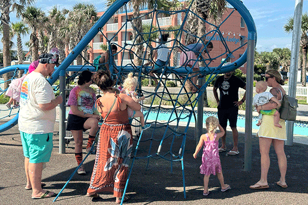 Adults stand by while their small children play on a climbing structure at a shaded park playground.