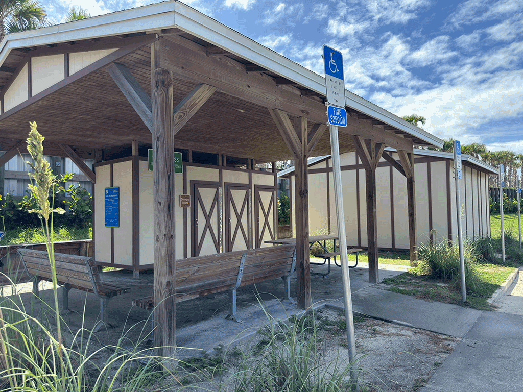 Changing rooms under a covered pavilion at a beachfront park, with benches and a picnic table.