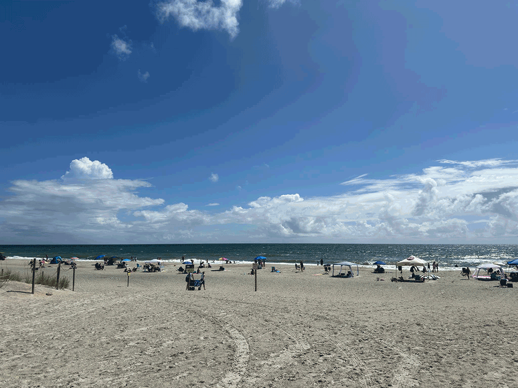 Many people walk or lounge on clear day at the beach, some under tents and umbrellas