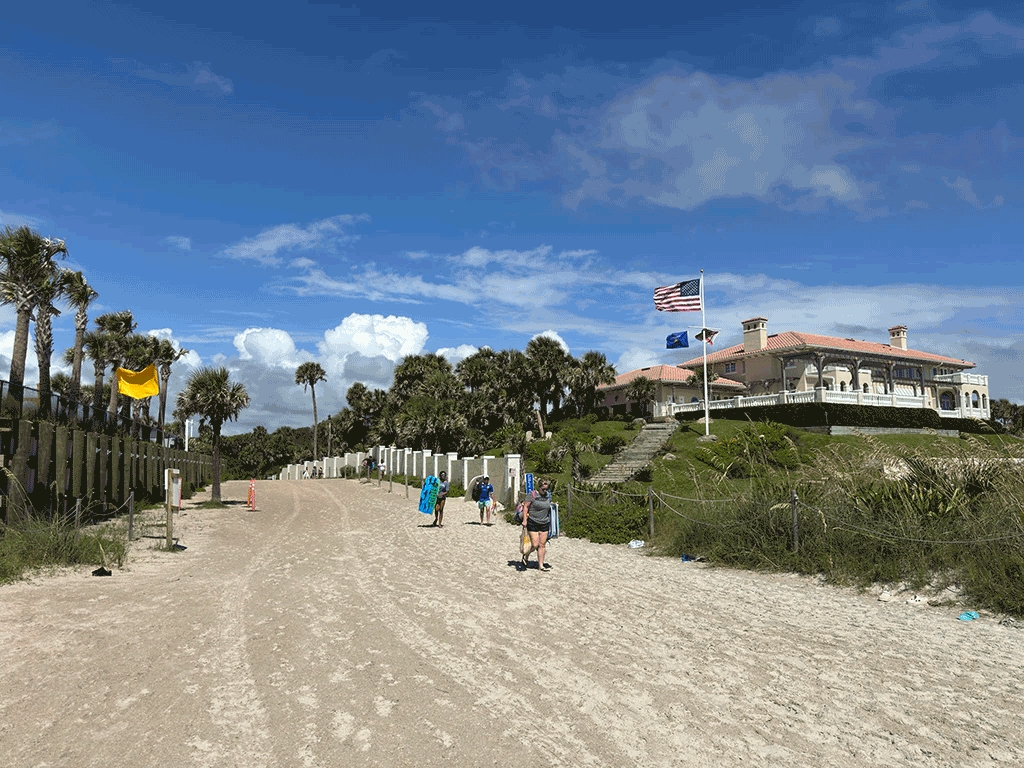 Several people walk down a beach access ramp toward the sand, with a clubhouse, palm trees, and waving flags in the background.