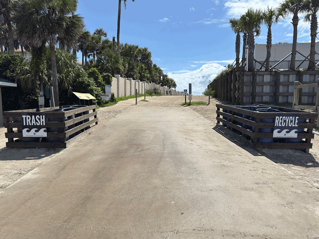 The beginning of a sandy vehicle access ramp toward a beach, flanked by bins marked ‘trash’ and ‘recycle.’