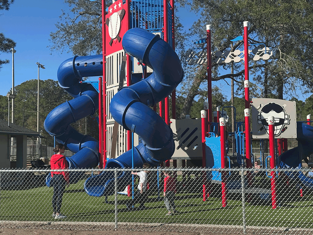 Moms watch their kids play on a baseball-themed play structure surrounded by a chain-link fence