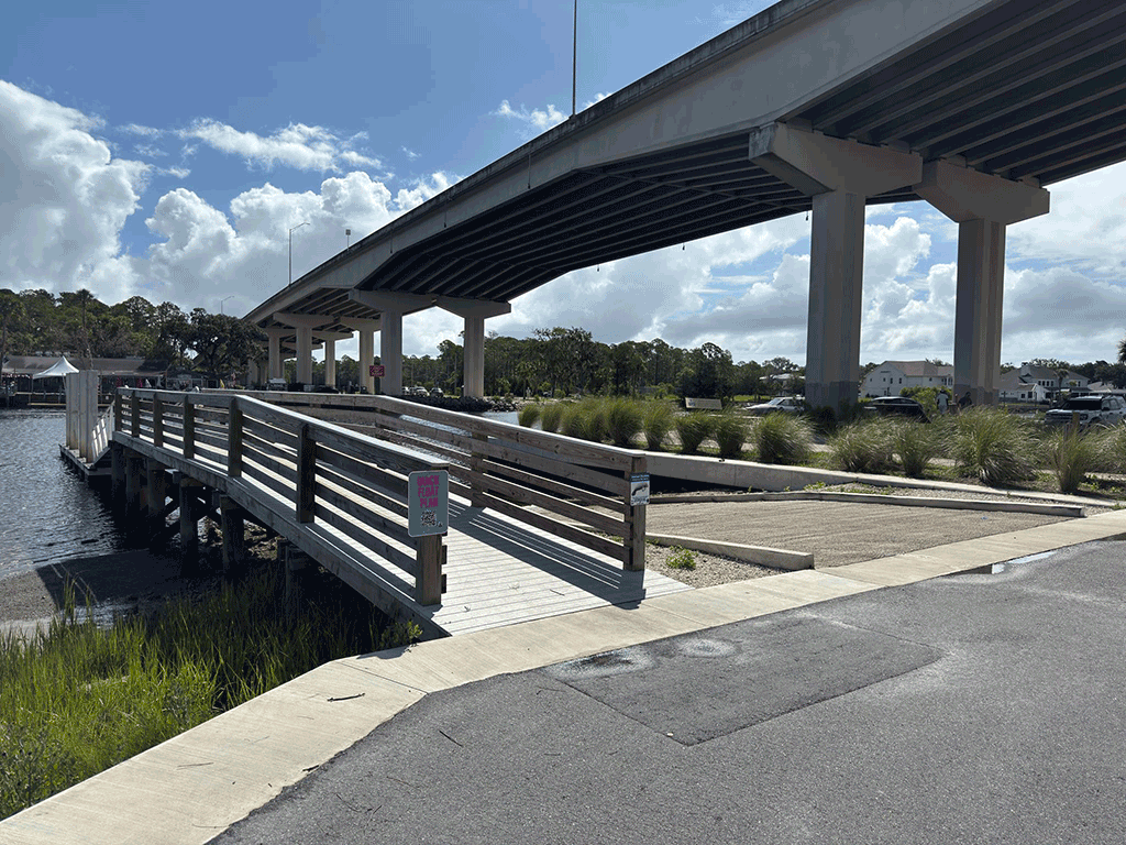 A wooden dock next to a boat ramp, with an overpass in the background.