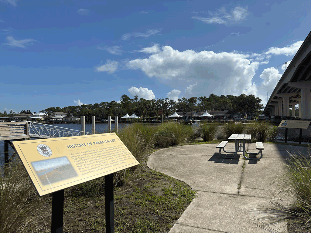A pair of historical signs by a picnic table, surrounded by tall grass, in between a boat ramp and an overpass.
