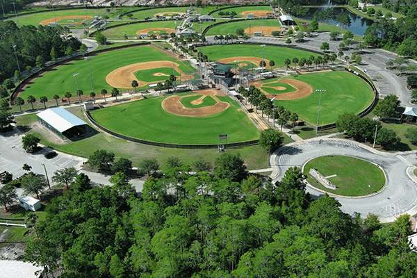 An aerial view of a sports complex with multiple baseball and softball fields, along with batting cages and parking spaces.