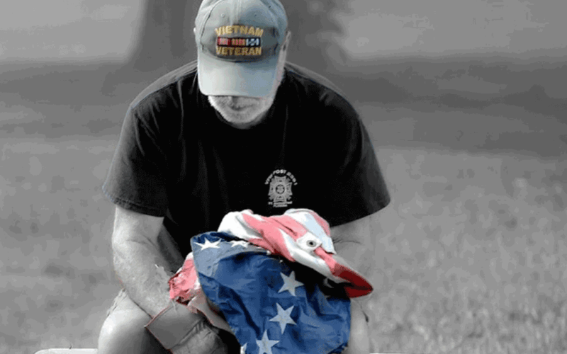 A veteran sits holding an American Flag. The image is without color except for the flag and his hat, reading Vietnam Veteran,