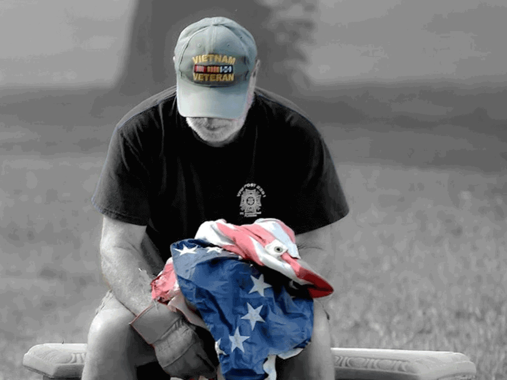 A veteran sits holding an American Flag. The image is without color except for the flag and his hat, reading Vietnam Veteran,