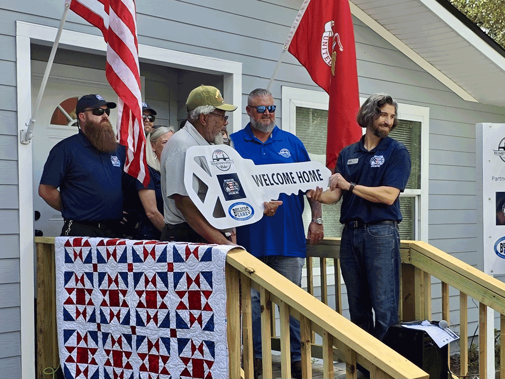 U.S. Marine Corps Vietnam War veteran Roy Wilbur stands with others on the front porch of a house holding a cardboard key that says "welcome home."