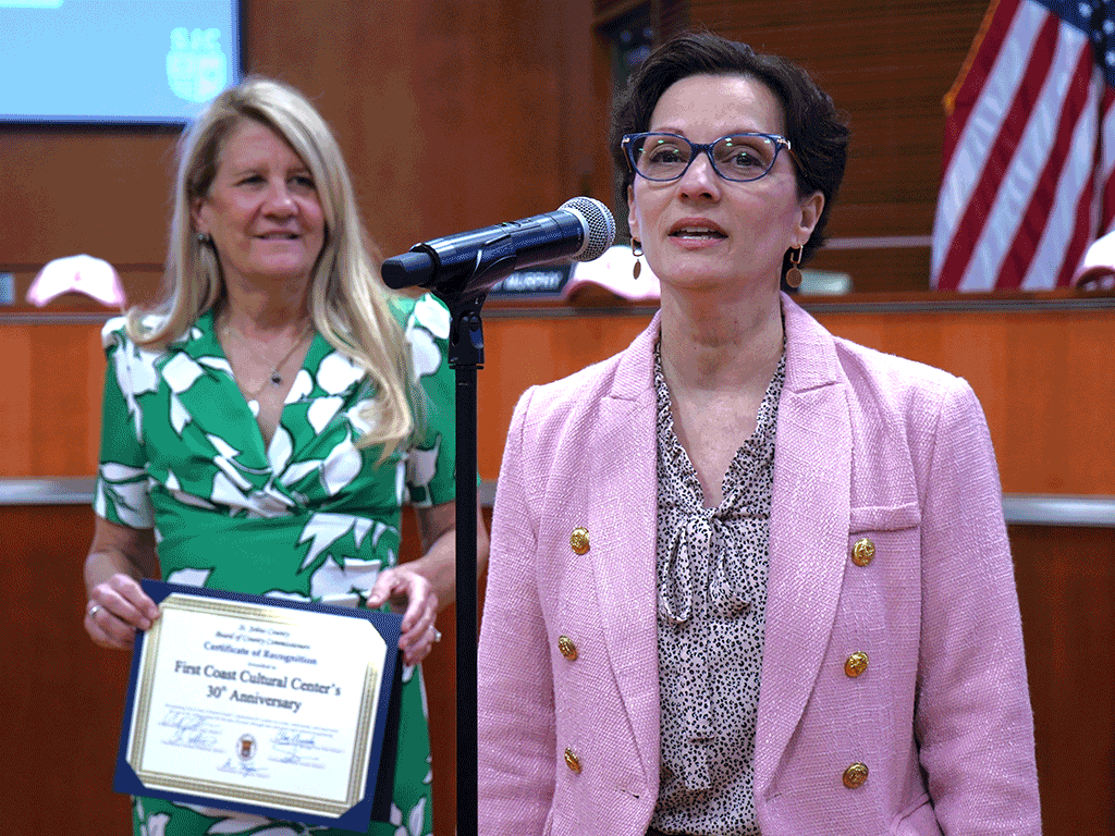 Commissioner Joseph stands with the certificate while Silvia Romero speaks into a microphone.