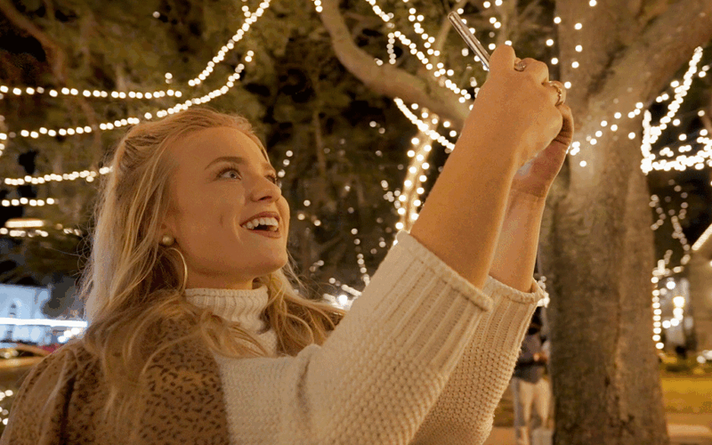 blonde woman holding phone up to take pictures of lighted holiday trees