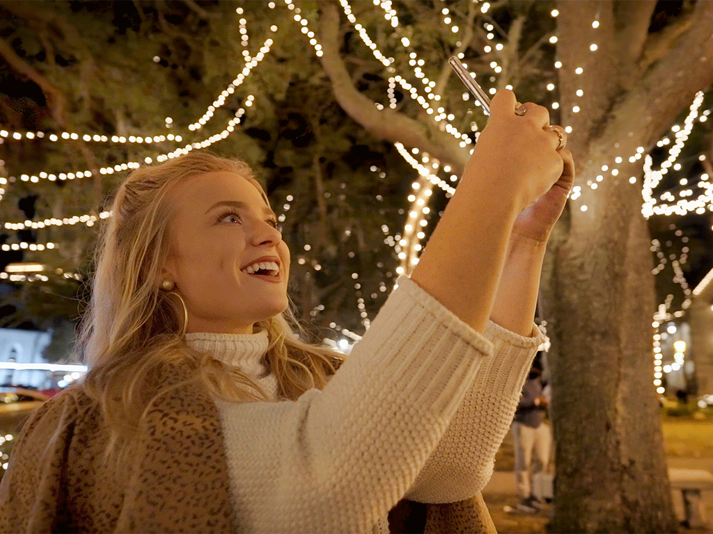blonde woman holding phone up to take pictures of lighted holiday trees